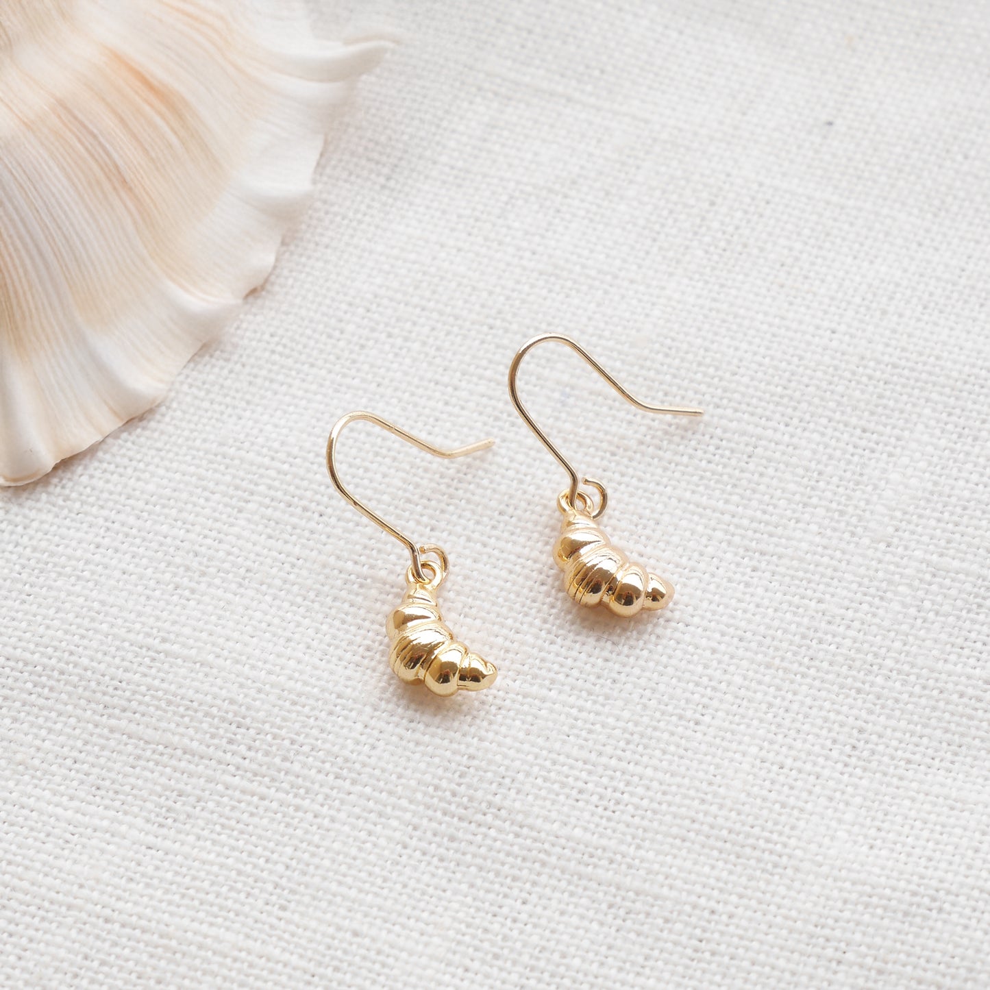 Gold croissant earrings on a textured white surface with a seashell in the background.