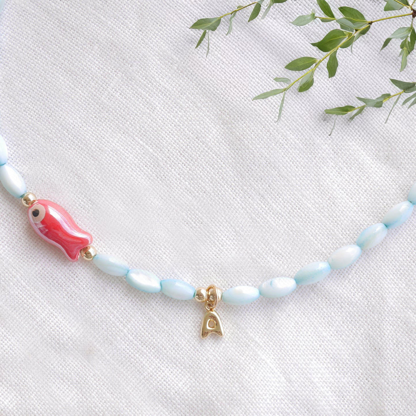 Turquoise beaded necklace with red fish and gold bubble initial charm, on a white cloth.