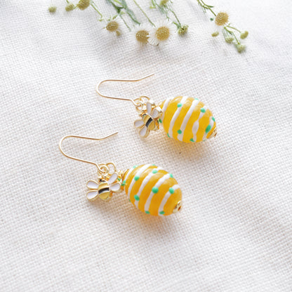 Yellow bee hive and bee charm gold dangle earrings, on a white cloth with some summer flowers.