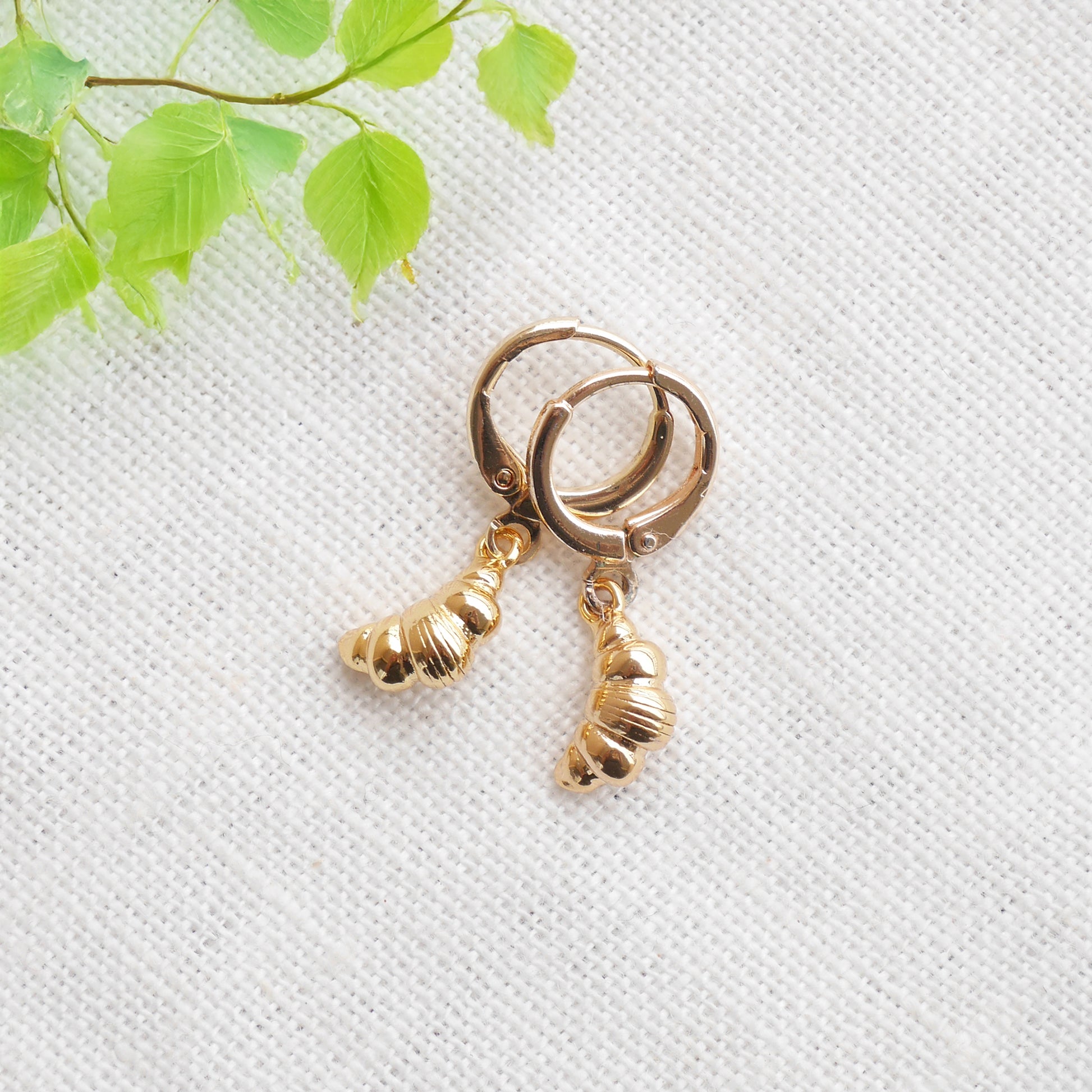 Gold croissant earrings on a textured white surface with a green leafy branch in the background.