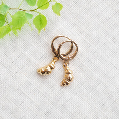 Gold croissant earrings on a textured white surface with a green leafy branch in the background.
