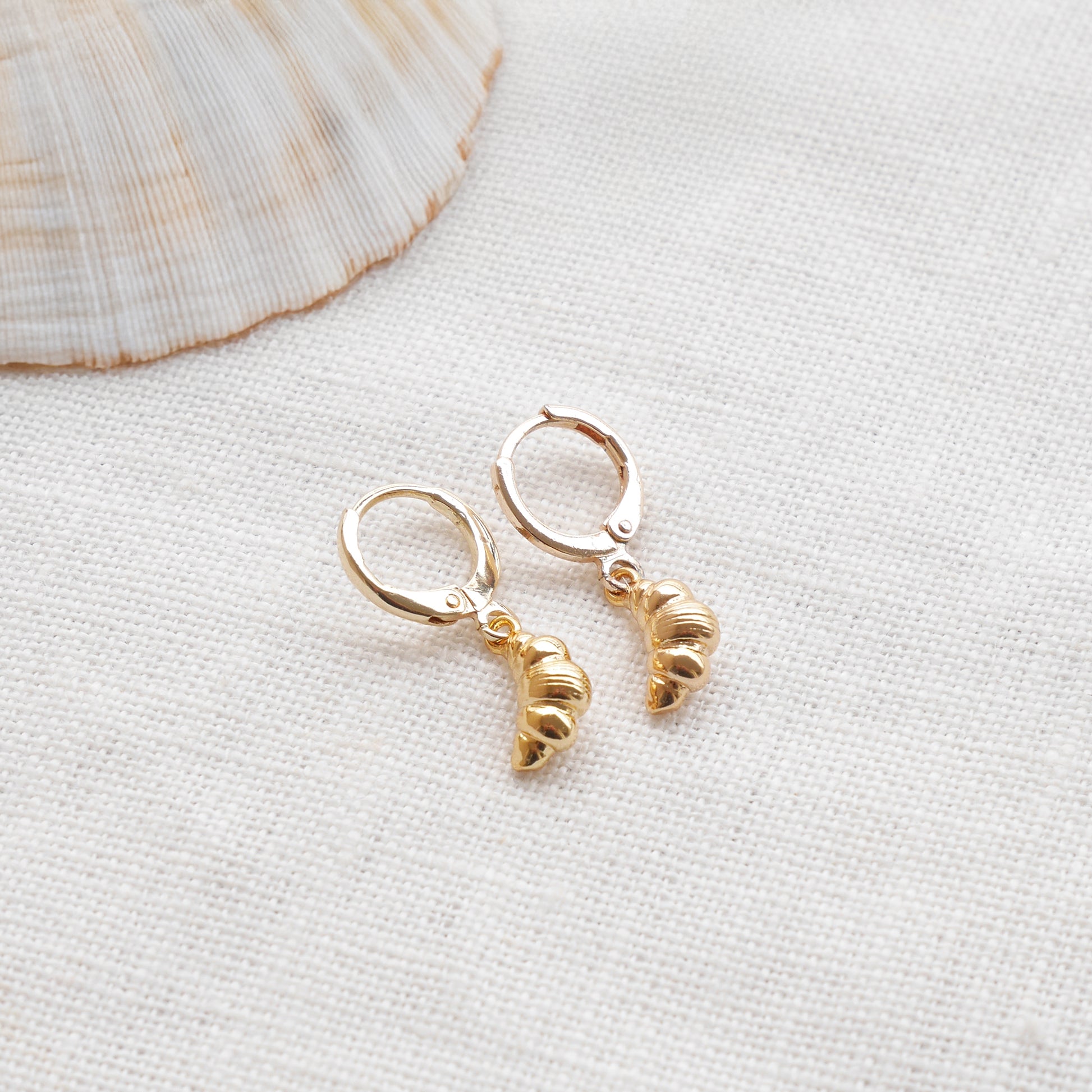 Gold croissant earrings on a textured white surface with a seashell in the background.
