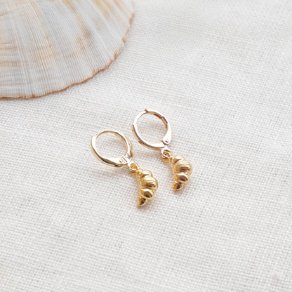 Gold croissant earrings on a textured white surface with a seashell in the background.