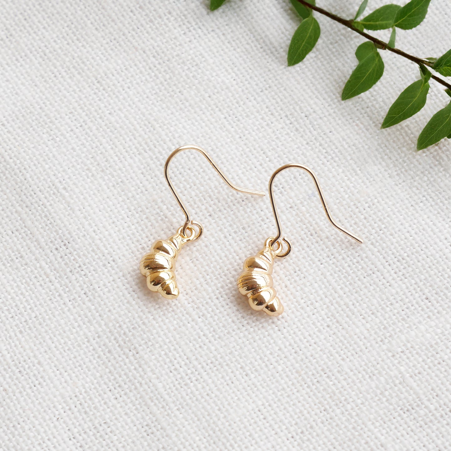 Gold croissant earrings on a textured white surface with a green leafy branch in the background.
