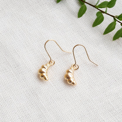 Gold croissant earrings on a textured white surface with a green leafy branch in the background.
