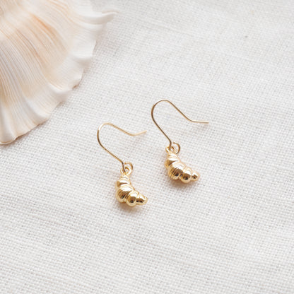 Gold croissant earrings on a textured white surface with a seashell in the background.