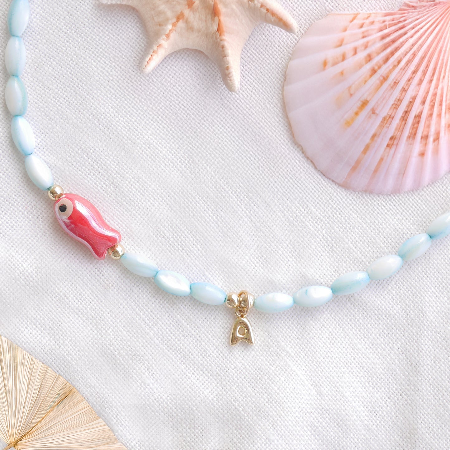Turquoise beaded necklace with red fish and gold bubble initial charm, on a white cloth.