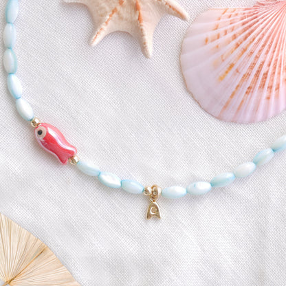 Turquoise beaded necklace with red fish and gold bubble initial charm, on a white cloth.