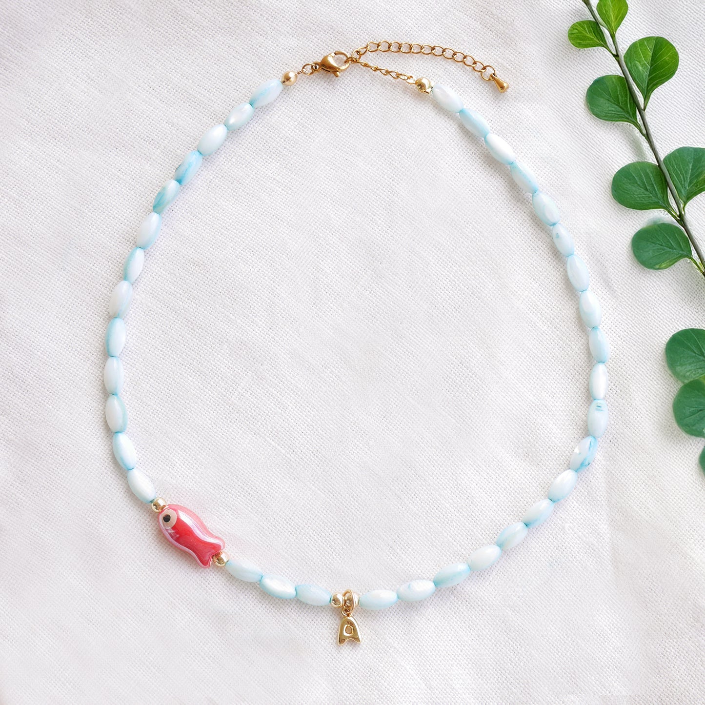 Turquoise beaded necklace with red fish and gold bubble initial charm, on a white cloth.