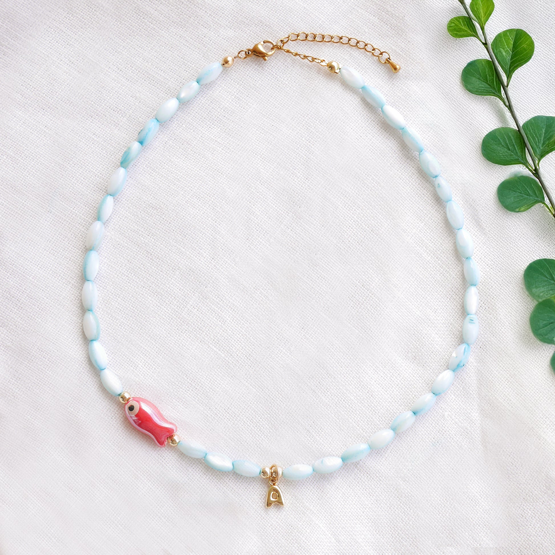 Turquoise beaded necklace with red fish and gold bubble initial charm, on a white cloth.