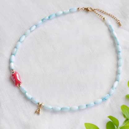 Turquoise beaded necklace with red fish and gold bubble initial charm, on a white cloth.