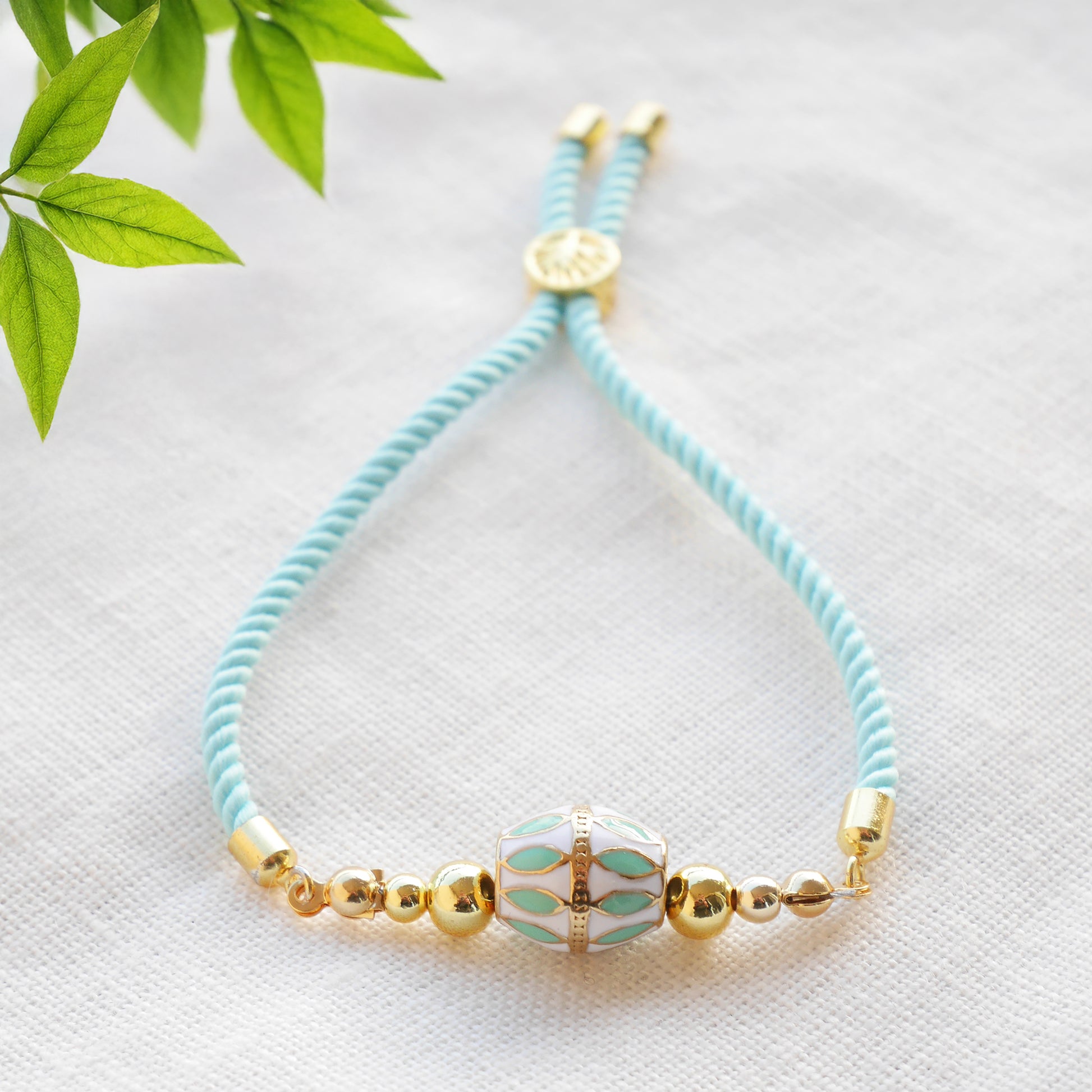 Turquoise adjustable nylon cord bracelet with 14k gold plated beads on a white cloth with a green leafy branch.
