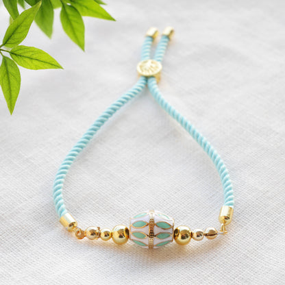 Turquoise adjustable nylon cord bracelet with 14k gold plated beads on a white cloth with a green leafy branch.