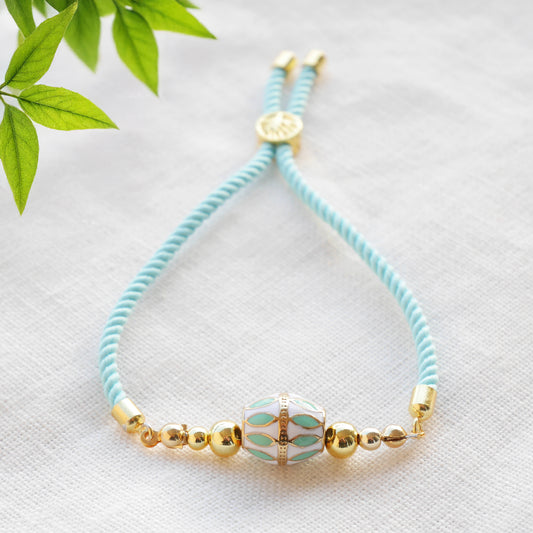 Turquoise adjustable nylon cord bracelet with 14k gold plated beads on a white cloth with a green leafy branch.