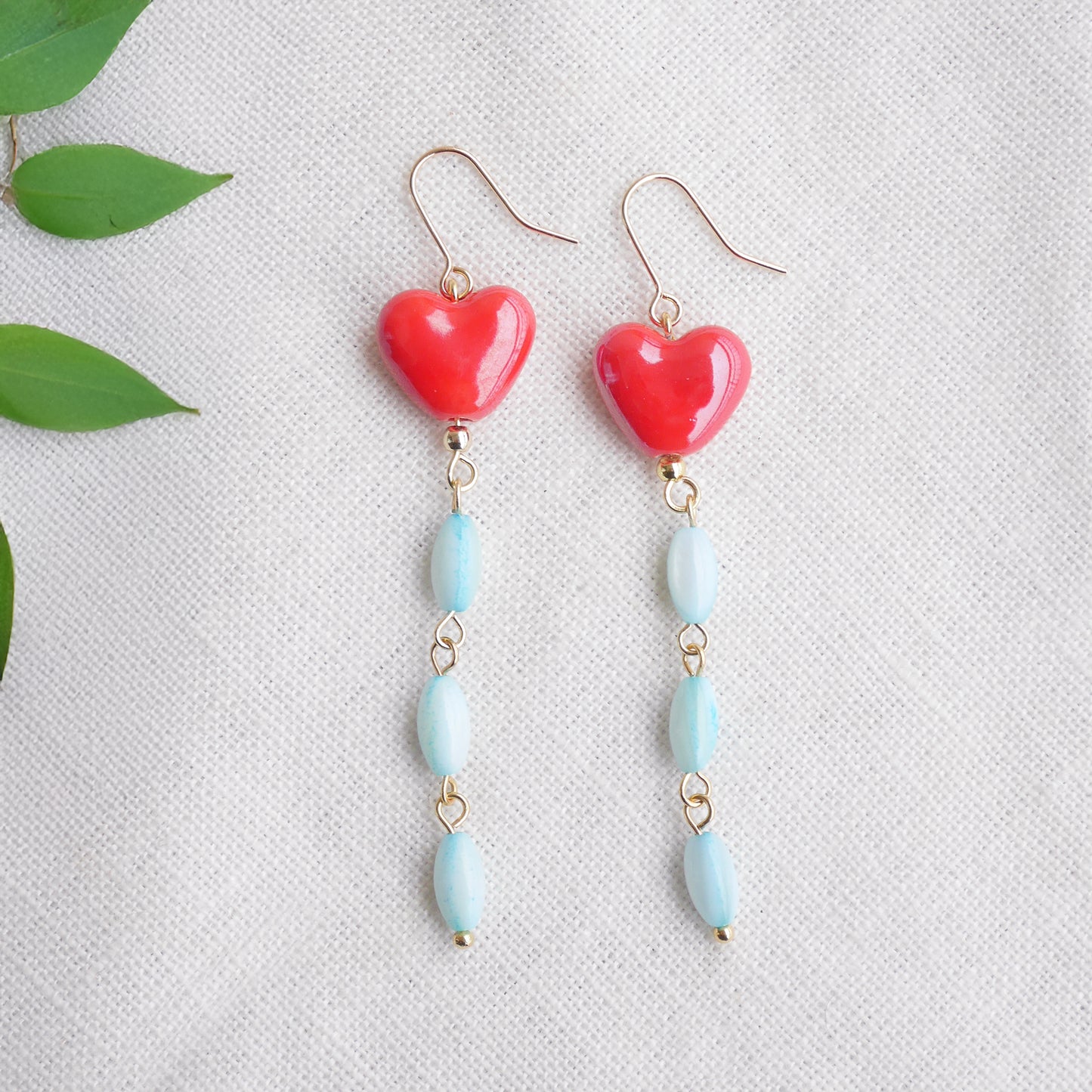 Long dangle earrings with red porcelain hearts and turquoise shell beads, on a white cloth.
