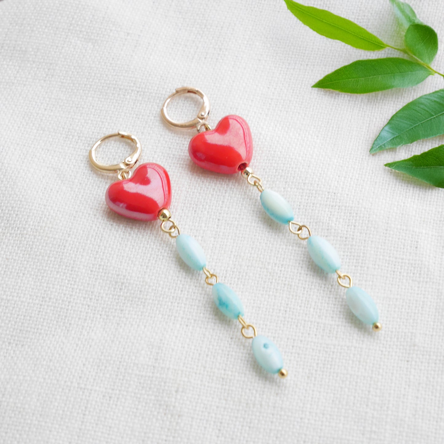 Long dangle earrings with red porcelain hearts and turquoise shell beads, on a white cloth.