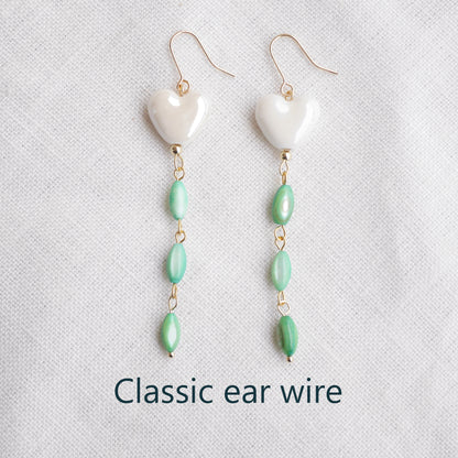 Pair of heart-shaped earrings with white porcelain heart and green shell beads on a light background.