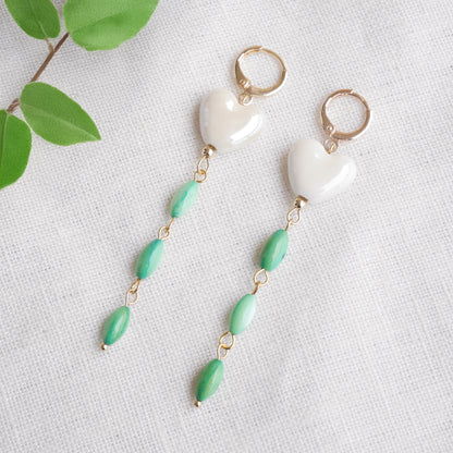 Pair of heart-shaped earrings with white porcelain heart and green shell beads on a light background.