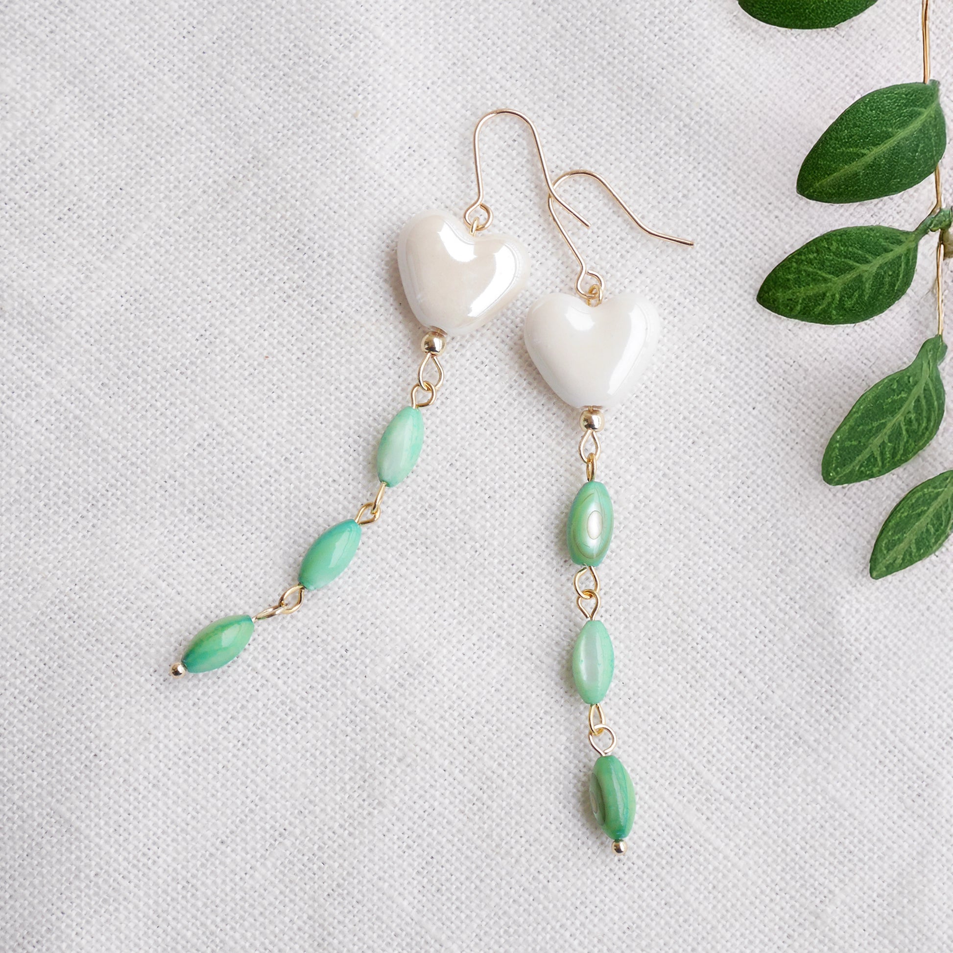 Pair of heart-shaped earrings with white porcelain heart and green shell beads on a light background.