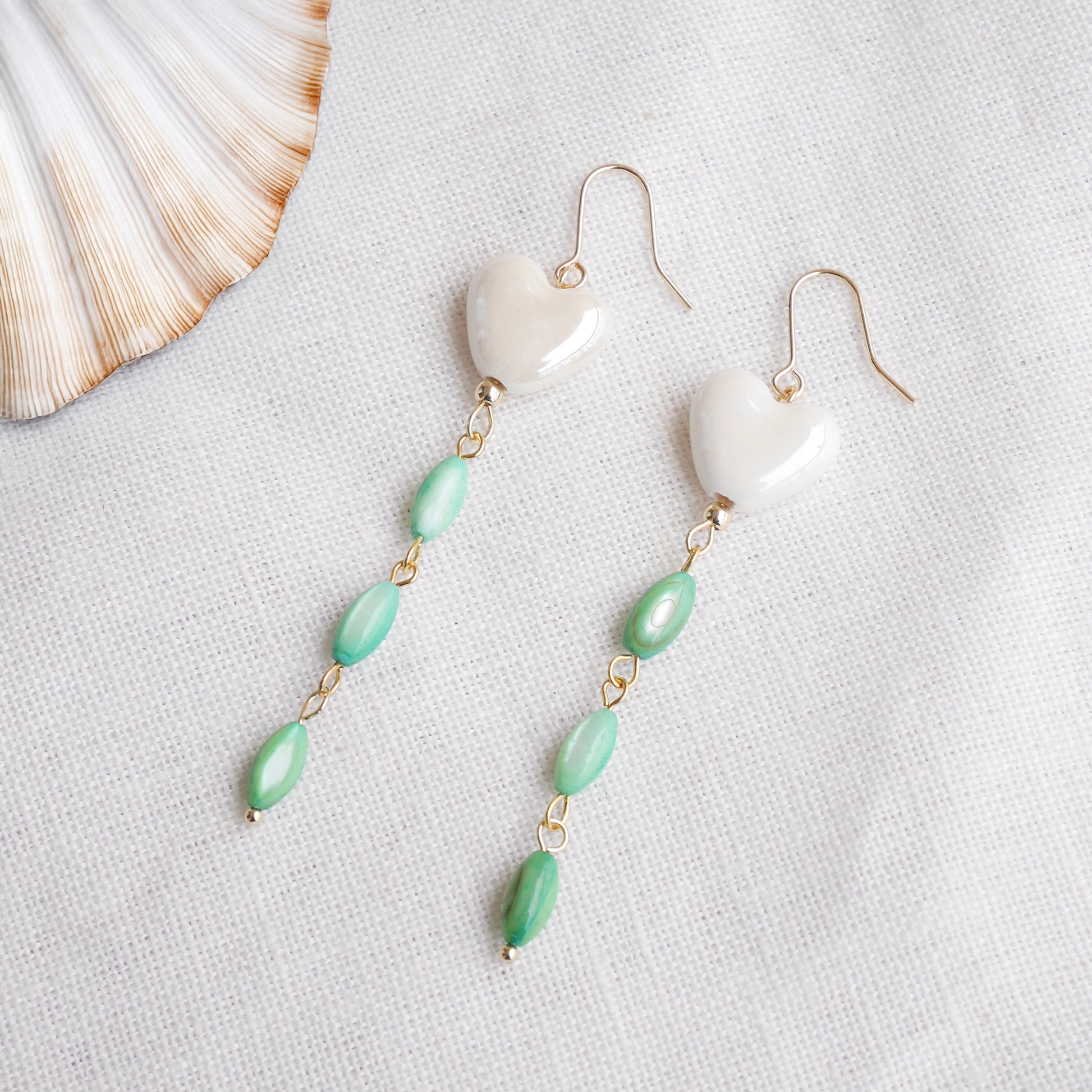 Pair of heart-shaped earrings with white porcelain heart and green shell beads on a light background.