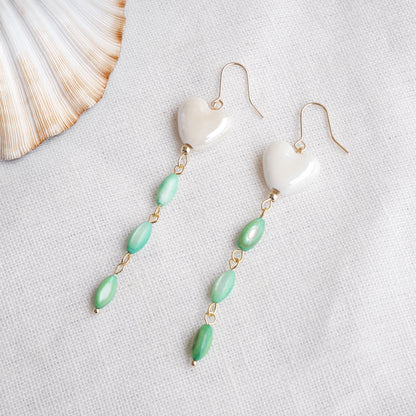 Pair of heart-shaped earrings with white porcelain heart and green shell beads on a light background.