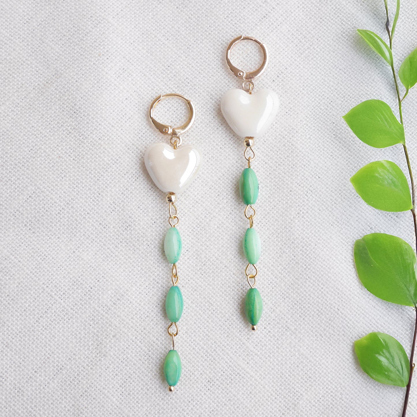 Pair of heart-shaped earrings with white porcelain heart and green shell beads on a light background